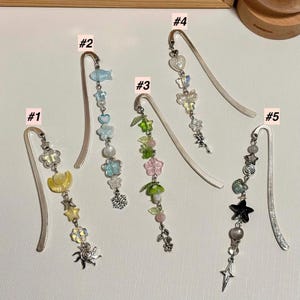 Puede incluir: Cinco marcadores de metal decorativos con colgantes. Cada marcador presenta un dise&ntilde;o diferente, incluyendo flores, estrellas y un sol. Los colgantes son de varios colores, como amarillo, azul y verde.