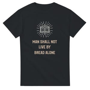 Op de afbeelding: Zwart T-shirt met een witte afbeelding van een open boek met een kruis en stralende lijnen. De tekst eronder luidt: "DE MENS LEEFT NIET VAN BROOD ALLEEN".