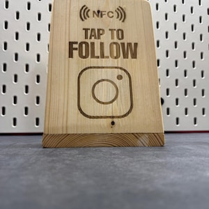 Könnte beinhalten: Ein Holzschild mit dem Instagram-Logo und dem eingravierten Schriftzug "TAP TO FOLLOW". Das Schild zeigt auch das NFC-Symbol. Das Schild ist aus hellem Holz gefertigt.