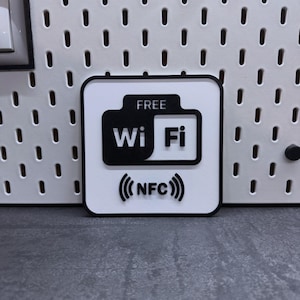 Könnte beinhalten: Ein quadratisches, weißes und schwarzes Wi-Fi-Schild mit den Worten "FREE Wi Fi" und "(NFC)". Das Schild hat einen schwarzen Rand und befindet sich auf einer grauen Oberfläche.