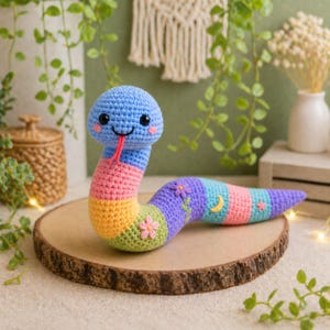 Puede incluir: Un juguete de serpiente de ganchillo colorido con cabeza azul, mejillas rosas y lengua roja. El cuerpo presenta rayas rosas, amarillas, verdes y moradas, con flores bordadas y una luna creciente. La serpiente está sobre una rodaja de madera.