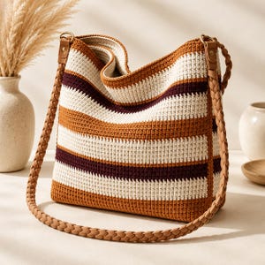 May include: A crochet hobo bag featuring horizontal stripes in cream, burnt orange, and deep maroon. The bag has a structured opening and a thick, braided tan shoulder strap attached with gold-toned metal rings on each side of the bag.