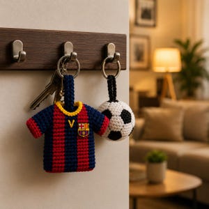 May include: Two handmade keychains hang from a wooden hook rack. One is a miniature crochet soccer jersey in blue, red, and yellow. The other is a crochet soccer ball in black and white. Keys are also hanging on the rack.