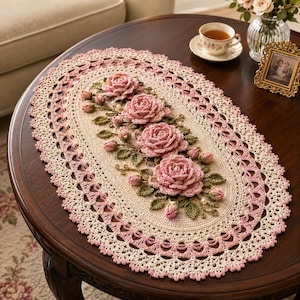 May include: An oval-shaped crochet doily featuring a cream-colored base with a decorative pink and cream scalloped border. The center is adorned with four three-dimensional crocheted pink roses, green leaves, and small white pearl accents.