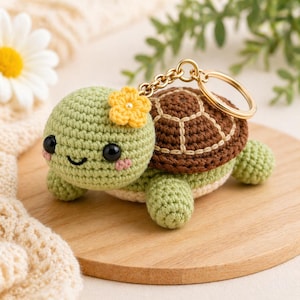 Può includere: Un portachiavi a forma di tartaruga all'uncinetto nei toni del verde, marrone e crema. La tartaruga ha una faccia sorridente, un fiore giallo e un anello portachiavi dorato. Il guscio della tartaruga è marrone con dettagli color crema.