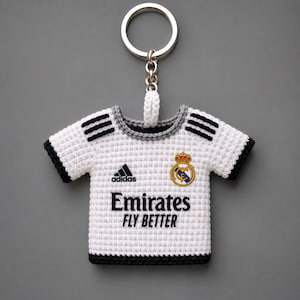Può includere: Portachiavi all'uncinetto bianco a forma di maglia da calcio. Presenta dettagli neri, tra cui strisce e finiture. Il logo Adidas, il testo Emirates Fly Better e uno stemma della squadra sono visibili. Anello portachiavi argentato.
