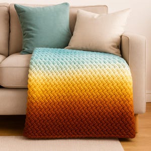 May include: A rectangular crochet blanket featuring a basketweave texture and a gradient color transition that shifts from a light teal at the top through yellow and orange to a deep rust brown at the bottom edge, draped over a beige sofa.
