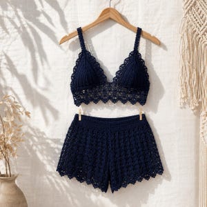 May include: A navy blue crochet two-piece set, including a triangle-shaped bralette and matching shorts. The bralette has adjustable straps and a scalloped edge. The shorts feature a similar openwork design and a scalloped hemline. The set is displayed on a wooden hanger.