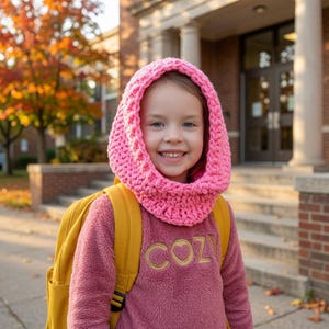 May include: A bright pink chunky knit hooded cowl worn over a mauve long-sleeve fleece pullover with the word COZY embroidered in gold thread on the chest, paired with a mustard yellow backpack, featuring a textured stitch pattern throughout.