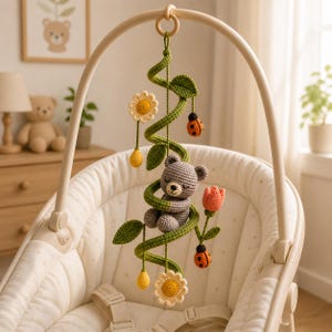 Könnte beinhalten: Handgefertigtes Baby-Mobile aus Häkelarbeit mit einem grauen Teddybären, der sich in einer grünen Ranke mit Blättern windet. Das Mobile enthält gelbe Blumen, Marienkäfer und eine orangefarbene Tulpe. Es hängt über einer cremefarbenen Babywippe.