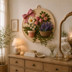 May include: A round, beige crochet wall hanging with a pink bow, daisies, tulips, and lavender. The flowers are in crochet pots and baskets. The wall hanging is displayed on a white wall above a wooden dresser.
