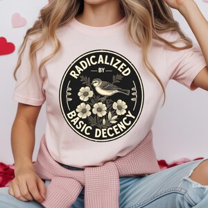 May include: A light pink t-shirt featuring a black circular graphic with the words "RADICALIZED BY BASIC DECENCY" surrounding a bird and floral illustration. The shirt is made of a soft material.