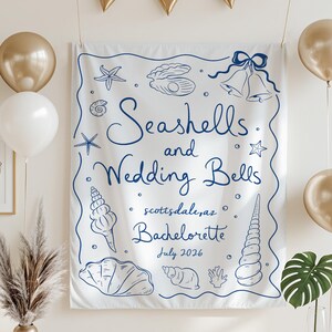 May include: White fabric banner with blue seashell and wedding bell illustrations. Text reads "Seashells and Wedding Bells, Scottsdale, AZ, Bachelorette, July 2026". Gold and white balloons and gold triangle banner decorate the background.