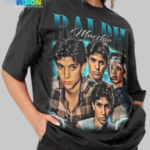 May include: Black unisex t-shirt featuring a graphic of Ralph Macchio with a blue and white design. The shirt has the text "RALPH Macchio" in a stylized font. The Fashion Fusion logo is in the upper left corner.
