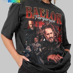 以下が含まれることがあります： 男性のグラフィックと赤い文字「BAELOR Targaryen」が特徴の黒いTシャツ。シャツはヴィンテージデザインで、左上にFashion Fusionのロゴがあります。