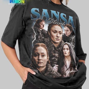 以下が含まれることがあります： ゲーム・オブ・スローンズのサンサ・スタークのグラフィックが特徴の黒いTシャツ。「SANSA Stark」という名前が青い文字で書かれ、キャラクターの複数の画像が含まれています。シャツの左上隅には「FASHION FUSION」のロゴがあります。