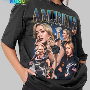 Puede incluir: Camiseta unisex negra con un collage de imágenes de Amber Glenn, con el nombre "AMBER Glenn" en letras doradas. La camiseta tiene el logo "FASHION FUSION" en la esquina superior izquierda. La camiseta es talla 2XL.