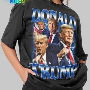 Puede incluir: Camiseta unisex negra con un gráfico de Donald Trump. La palabra "DONALD" está en azul encima de la imagen y "TRUMP" en azul debajo. La camiseta tiene el logo "FASHION FUSION" en la esquina superior izquierda.