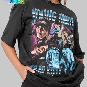 May include: Black oversized t-shirt featuring a graphic of a woman wearing a cap, with the text "YOUNG MIKO" and "TRAP KITTY" in a blue, retro style. The shirt has the "FASHION FUSION" logo at the top.