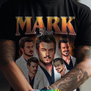 May include: Black t-shirt featuring a graphic of Mark Sloan with the name in gold and orange lettering. The shirt includes multiple images of the character. The bottom of the shirt has the text "FASHION FUSION" in a colorful gradient.