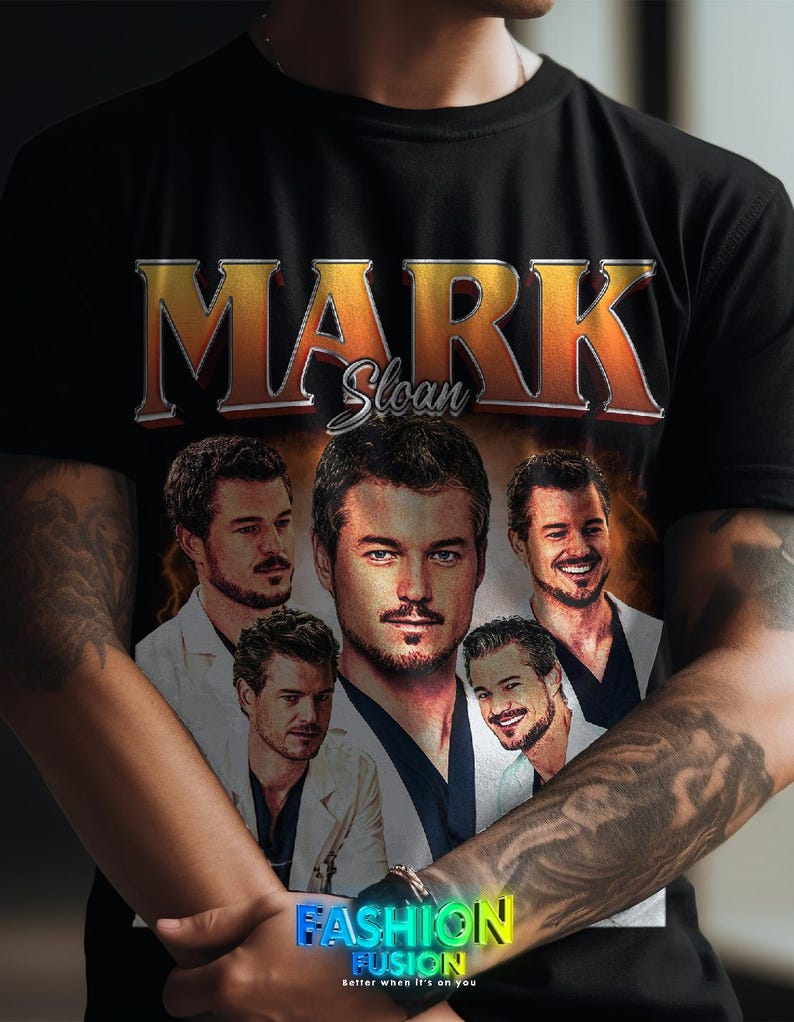 May include: Black t-shirt featuring a graphic of Mark Sloan with the name in gold and orange lettering. The shirt includes multiple images of the character. The bottom of the shirt has the text "FASHION FUSION" in a colorful gradient.