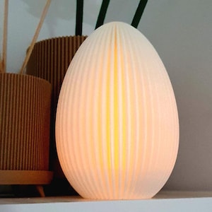 Könnte beinhalten: Eine weiße, eiförmige Lampe mit vertikalen Rippendetails. Die Lampe strahlt ein warmes, gelbes Licht aus. Im Hintergrund befinden sich zwei braune, zylindrische Behälter mit vertikalen Rippendetails sowie Holzstäbchen.