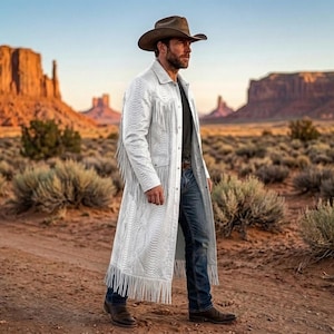 Puede incluir: Un abrigo largo blanco con un patrón de piel de serpiente texturizado, con flecos largos en el pecho, las mangas y el dobladillo. El abrigo se lleva sobre una camisa negra y vaqueros azules, junto con un sombrero de vaquero marrón y botas de cuero marrón.