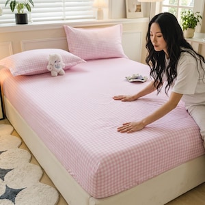 May include: A bed set featuring a fitted sheet and two pillowcases with a light pink and white gingham check pattern. A small white plush toy sits on one pillowcase, and the bedding is spread over a mattress on a white bed frame.