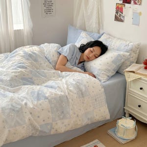 May include: A light blue and white patchwork bedding set featuring a pattern of small flowers, stars, and bows. The set includes a duvet cover and matching pillowcases, displayed on a bed with a solid light blue fitted sheet and a light blue pajama top.