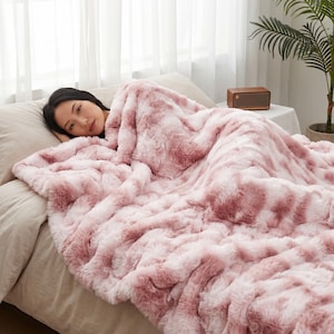 Può includere: Una coperta in pelliccia sintetica felpata in una palette di colori variegati rosa antico e bianco. La coperta presenta una consistenza spessa e arruffata con fibre a pelo lungo che creano un aspetto morbido e stratificato sul letto.