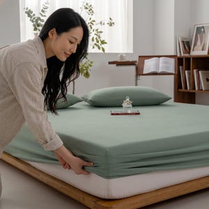 Puede incluir: Una sábana bajera de color verde salvia que cubre un colchón sobre una estructura de cama baja de madera. La sábana es de un color verde sólido y apagado con una textura suave, tensada sobre las esquinas del colchón, junto a dos fundas de almohada a juego.