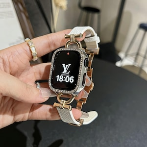 May include: A silver smartwatch with a white leather strap and gold-tone accents. The watch face displays the time 18:06 and a logo. The bezel is encrusted with small, sparkling stones. A matching ring is also visible.