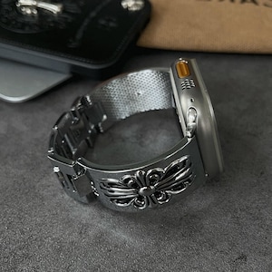 Peut inclure: Montre connectée argentée avec un bracelet décoratif orné d'un motif floral. La montre a un bracelet en maille et un cadran rectangulaire avec un accent jaune. La montre est présentée sur une surface grise.