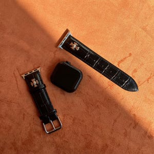 Peut inclure: Bracelets de montre Apple Watch en cuir noir avec des éléments argentés et un motif de croix décoratif. Les bracelets sont présentés avec un boîtier de montre noir sur une surface orange.