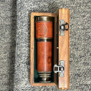 Puede incluir: Un telescopio vintage de latón y cuero en un estuche de madera. El telescopio es de cuero marrón con detalles de latón y el estuche es de madera marrón claro. El estuche está abierto, revelando el telescopio en su interior. La parte superior del telescopio tiene la palabra "LONDON".