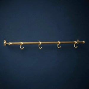 May include: A horizontal wall-mounted rail made of brushed brass metal, featuring four matching sliding hooks. The rail is secured at both ends with rectangular mounting plates, each containing two screw holes for attachment to a flat surface.