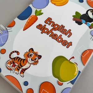 May include: Spiral-bound English alphabet learning book. The cover features a cartoon tiger, a carrot, a banana, an apple, and a toucan. The words "English alphabet" are printed in orange. The background has colourful shapes.
