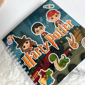 Può includere: Un quaderno a spirale con un'illustrazione colorata di tre personaggi dei cartoni animati, un treno rosso, un serpente verde, un boccino d'oro e uno stemma. Il testo Harry Potter è stampato in grandi lettere arancioni sulla copertina.