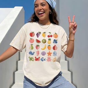 May include: A cream-colored short-sleeve t-shirt featuring a grid of colorful, patterned illustrations of various fruits and sea creatures, including an apple, grapes, watermelon, pineapple, whale, crab, and starfish, arranged in rows.
