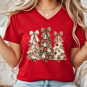 May include: A red V-neck t-shirt featuring a graphic print of three stylized Christmas trees. Each tree is decorated with different patterns including plaid, leopard print, and floral motifs, topped with large bows and small gold stars.