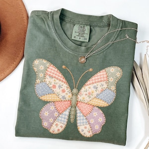 May include: An olive green short-sleeve t-shirt featuring a large butterfly graphic on the front. The butterfly is composed of various patchwork-style fabric patterns including floral, gingham, and polka dot prints in soft pastel colors like pink, blue, and yellow.