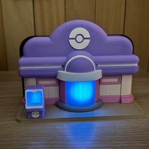 Könnte beinhalten: Ein lila-weißes Gebäudemodell mit einem blau leuchtenden Mittelteil. Das Modell hat eine abgerundete Oberseite mit einem weißen Emblem. Links befindet sich eine kleinere passende Struktur. Die Basis ist hellbraun.