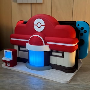 Könnte beinhalten: Eine rot-weiße, 3D-gedruckte Ladestation in Form eines Gebäudes, mit blauem Licht, das aus der Mitte kommt. Ein blauer und roter Gamecontroller ist an der Station befestigt. Auf der linken Seite befindet sich ein kleiner roter Spielautomat.