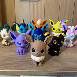 May include: A collection of nine small, colorful plastic figurines representing various fictional creatures. The group includes characters in shades of brown, purple, blue, orange, yellow, black, and pink, each featuring distinct ear shapes and facial markings.