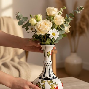 Può includere: Un vaso di ceramica bianco con un motivo floreale, che contiene un bouquet di rose color crema, margherite ed eucalipto. Il vaso presenta un motivo a righe bianche e nere e un motivo floreale rosso e verde.