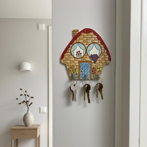 May include: A house-shaped key holder made of wood, featuring a red roof, brick-effect walls, and a blue door. It has three hooks with keys attached. The design includes painted flowers and two round windows with curtains.