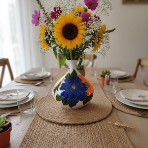 Könnte beinhalten: Ein Esstisch mit einem Jute-Tischläufer und Platzsets. Eine weiße Vase mit blauen und orangefarbenen Blumendesigns enthält einen Strauß Sonnenblumen, rosa Kosmeen und Schleierkraut. Teller, Gläser und Besteck sind für eine Mahlzeit gedeckt.
