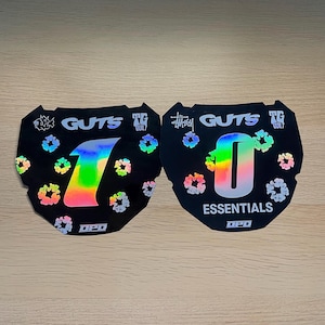 May include: Two black motorcycle number plate decals with holographic accents. The left decal features the number 1, while the right features the number 0 and the text ESSENTIALS. Both include GUTS, TG BUILT, and OPD logos with small floral patterns.