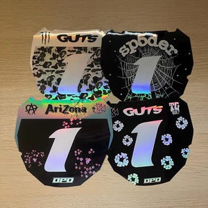 Peut inclure: Quatre autocollants de plaque d'immatriculation de moto affichant le chiffre 1 dans une grande police holographique centrale. Les motifs incluent un camouflage, une toile d'araignée, des fleurs de cerisier avec AriZona et des motifs floraux.
