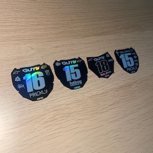 Puede incluir: Cuatro pegatinas negras e iridiscentes con números y texto blancos. Las pegatinas muestran los números 15, 16 y 18, junto con la palabra "GUTS" y otro texto. Las pegatinas tienen varios logotipos y la palabra "PRICKLY".